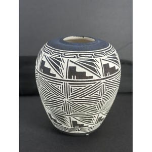 Puede incluir: Jarrón de cerámica redondo con borde azul oscuro y un patrón geométrico en blanco y negro. El jarrón presenta un diseño repetitivo de formas y líneas angulares, creando un contraste visual llamativo. Altura aproximada 15 cm.