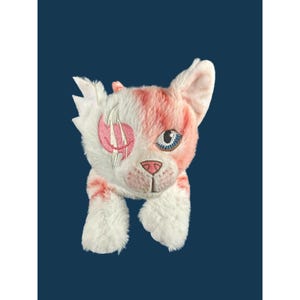 May include: A plush cat toy featuring white and pink fur. The cat has one blue eye and one pink embroidered eye patch with white claw marks. It has a pink nose, stitched whiskers, and pointed ears. The plush is soft and textured throughout.