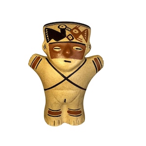 Puede incluir: Una figurita de cerámica pintada a mano que representa a una persona con un tocado marrón y blanco y un cuerpo marrón y blanco. La figurita está de pie con los brazos extendidos.