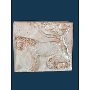 Puede incluir: Un azulejo de cerámica cuadrado que presenta una escultura en relieve de un caballo de pie junto a un potro más pequeño. La pieza tiene una base de arcilla color terracota con acentos de esmalte blanco que resaltan las figuras y un diseño de rama de árbol en el fondo.
