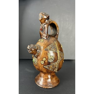 May include: A brown ceramic vessel with a rounded body and a pedestal base. The piece features two sculpted animal heads, one at the top and one on the side, decorated with gold-toned metal accents, small green and red stones, and a llama motif.