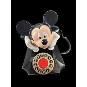 May include: A vintage Mickey Mouse telephone in black, white, and tan. The phone features Mickey's face as the receiver, with his ears as the top of the phone. The rotary dial is gold with a red center.