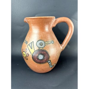May include: A terracotta pitcher with a brown, yellow, and blue geometric design. The pitcher has a wide, round body and a handle.