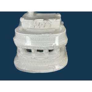 Puede incluir: Modelo de cerámica blanca de un restaurante drive-in, envuelto en plástico transparente. El texto "Flo's" es visible en la parte superior y "Drive-In" en el lateral. El modelo tiene un diseño arquitectónico detallado.