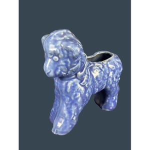 以下が含まれることがあります： 青いセラミックプードルの置物。表面はテクスチャ加工が施されています。プードルは首に装飾用のリボンがあり、背中はくり抜かれており、プランターや花瓶として使用できる可能性があります。置物は単色の青色です。