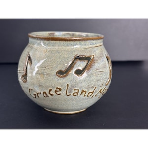 May include: A light blue ceramic candle holder with cut-out music notes and the words "Grace Land, Music" in brown glaze.