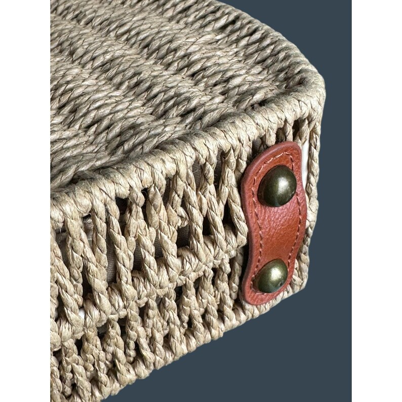 May include: Close-up of a woven straw bag with a rounded shape. The bag is made of natural-colored straw and features a brown leather accent with two brass-colored rivets. The weave is intricate and detailed.