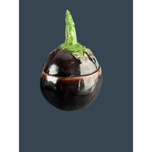 May include: A ceramic eggplant-shaped container with a lid. The container is a deep purple color with a brown rim and a green stem. The lid fits snugly on the container. The design is a whimsical and decorative piece.