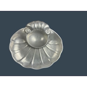 May include: A silver-colored, shell-shaped dish with a central, rounded depression. The dish has a textured surface with radiating lines and decorative swirl details. The overall design is reminiscent of a scallop shell, suitable for serving dips or small items.