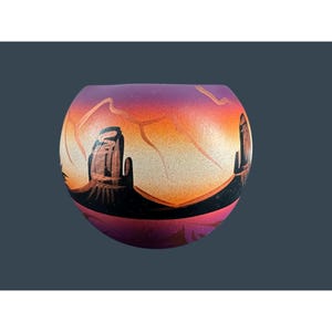 May include: A round, hand-painted decorative bowl featuring a desert landscape scene. The bowl has a gradient of colors, including purple, orange, and black. The design depicts mesas and a sunset.