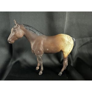 Pode incluir: Uma figura de cavalo marrom e branco com manchas, com uma crina e cauda pretas. A figura está em pé sobre uma superfície preta.