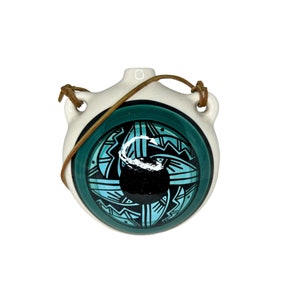 Może przedstawiać: Biała ceramiczna piersiówka z dekoracyjnym turkusowo-czarnym geometrycznym wzorem. Piersiówka ma ciemnozieloną obwódkę i brązowy skórzany pasek. Piersiówka została zaprojektowana z dwoma otworami na pasek.