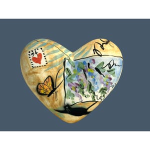 May include: Heart-shaped ceramic art piece with a painted design. The artwork features a butterfly, a postage stamp with a heart, and a floral scene. The color palette includes beige, blue, purple, and black.