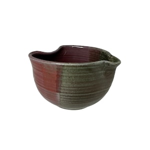 Puede incluir: Un bol mezclador de cerámica verde y morado con un pico vertedor único, ligeramente descentrado. El bol tiene un aspecto rústico y hecho a mano.