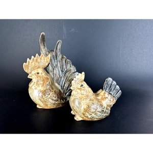 May include: A set of two ceramic rooster figurines. The larger rooster is brown and white with a black tail. The smaller rooster is brown and white with a black tail and a black head.