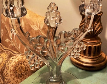May include: A clear glass three-arm candelabra with a floral design. The candelabra has a round base and three arms that curve upwards. Each arm has a cup-shaped holder at the top.