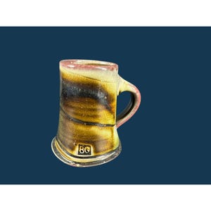 Puede incluir: Una taza de cerámica con un esmalte reactivo en tonos mezclados de amarillo mostaza, marrón oscuro y negro. La taza tiene una base ancha, un asa curva y un borde distintivo. Las letras BG están estampadas en un cuadrado cerca de la base.