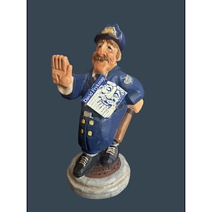 May include: A hand-carved wooden figurine of a police officer in a blue uniform and hat, holding a baton and a book labeled "David Frykman." The figurine stands on a round, stone-like base with a manhole cover design.