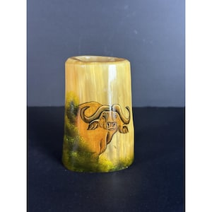 Könnte beinhalten: Eine kleine, zylindrische, handbemalte Holzvase mit einem braunen Büffelmotiv. Die Vase ist hellbraun mit einem grün-gelben Hintergrund.