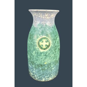 Puede incluir: Un jarrón de cerámica alto con un esmalte moteado verde y azul. El jarrón presenta un borde azul claro y un emblema circular con un diseño de cruz. El jarrón mide aproximadamente 25 cm de alto.
