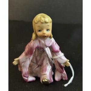 May include: A porcelain figurine of a young girl with blonde hair and blue eyes. She is wearing a pink and purple dress with petal-like details. The figurine has a gold-colored foot and is holding a white string.