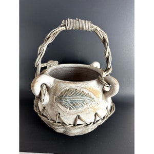 May include: A white ceramic planter with a woven wicker handle and a leaf design. The planter has a rustic, earthy look.