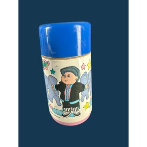 Op de afbeelding: Een cilindrische thermoskan met een felblauw deksel en een lichtroze onderkant. De witte behuizing is voorzien van een afbeelding van een stripfiguur met donker haar, gekleed in een zwart jasje, roze overhemd en gestreepte broek.
