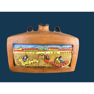 May include: A brown ceramic canteen with two handles and a central relief panel depicting a village scene with figures in colorful clothing, baskets of produce, and red-roofed buildings. The artist signature R. Lopez is visible on the panel.