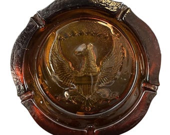 May include: Amber-colored glass ashtray with a detailed embossed image of an eagle with outstretched wings, a symbol of the United States. The ashtray has a scalloped edge and a circular design. The glass has a glossy finish.