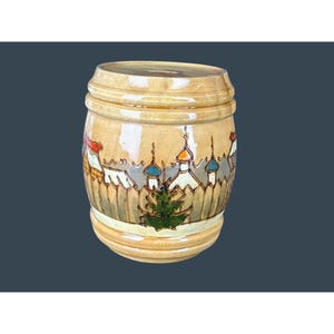 May include: A cylindrical, ceramic container with a lid, featuring a hand-painted village scene. The artwork includes buildings with colorful domes, a green fir tree, and a wooden fence. The container is tan with a glossy finish.