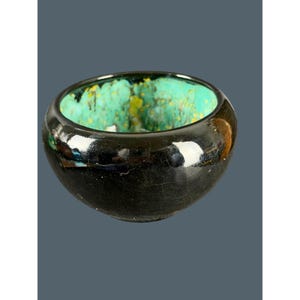 May include: A small, round ceramic bowl with a glossy black exterior featuring a fine crackle glaze pattern. The interior of the bowl is a vibrant, mottled turquoise green with scattered yellow and dark blue speckles throughout.