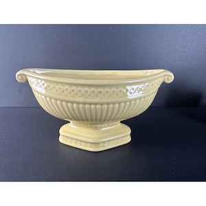 Puede incluir: Una jardinera de cerámica amarillo pálido con base de pedestal. La jardinera tiene una abertura ovalada ancha con asas enrolladas en cada extremo. Las crestas verticales adornan el cuerpo y un patrón decorativo rodea la parte superior.