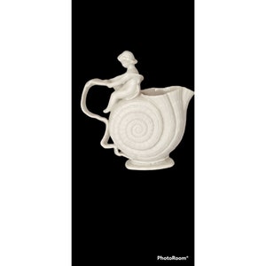 May include: White ceramic pitcher shaped like a seashell with a cherub figurine sitting on top. The pitcher has a spiral design and a handle.