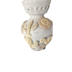May include: A small, off-white ceramic vase with a textured surface. The vase features relief details of cacti, a sun with a face, and other desert-themed elements. The vase has a decorative rim and the initials "C.O.I." are visible.