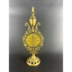 Puede incluir: Un frasco de perfume de metal dorado con un soporte decorativo. El frasco tiene un inserto redondo de vidrio amarillo con un diseño floral. El soporte tiene una parte superior redonda y tres patas.