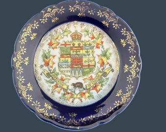 May include: A decorative plate with a dark blue rim and a central design featuring the Canadian coat of arms. The rim is adorned with gold floral patterns. The plate's center displays a detailed emblem with a beaver and the words "Dominion of Canada."