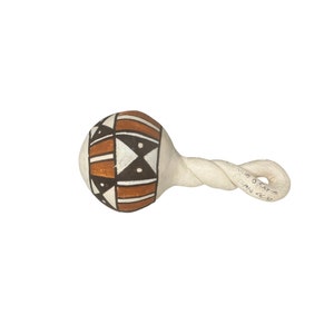 Puede incluir: Un sonajero pintado a mano con un mango blanco retorcido. La cabeza bulbosa del sonajero presenta un diseño geométrico en marrón, negro y blanco. El mango tiene texto escrito a mano. El sonajero está sobre un fondo blanco.