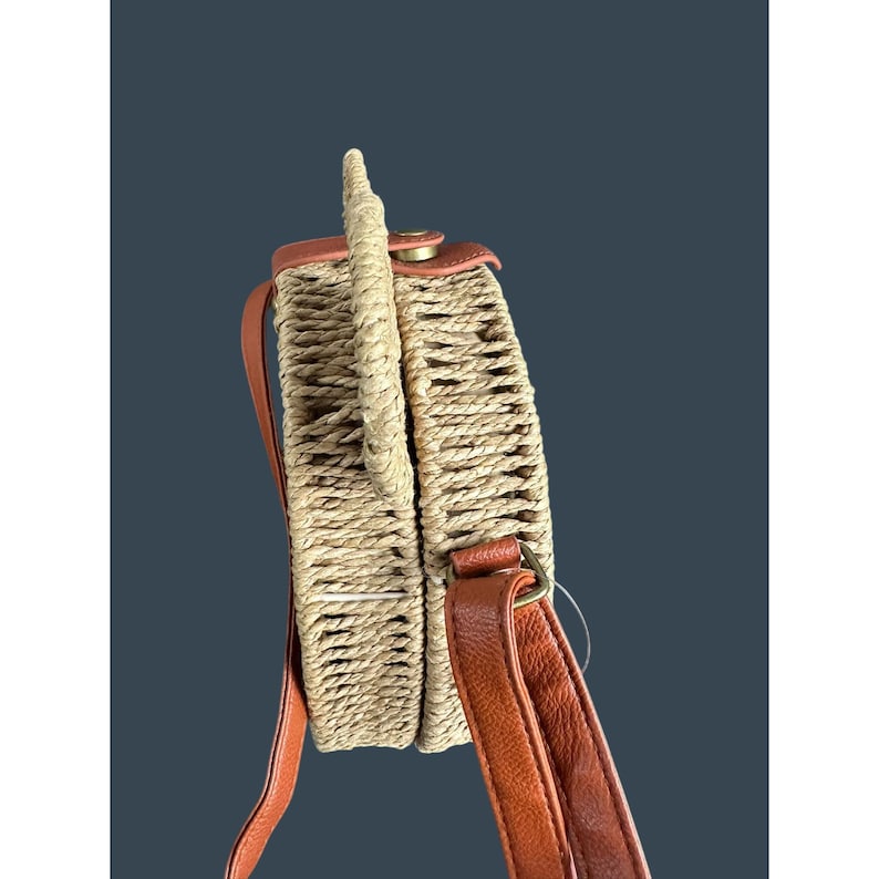 May include: A round, woven straw handbag with a brown leather handle and shoulder strap. The bag has a circular shape and a woven texture. The strap and handle are a warm brown color, and the bag is a natural straw color.