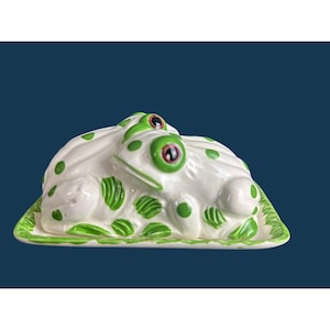May include: A white ceramic butter dish shaped like a frog with green spots and red eyes. The lid features a sculpted frog resting on a rectangular base with a green painted edge. The glossy finish highlights the smooth curves of the frog.
