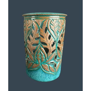 May include: A turquoise ceramic vase with a leaf design. The vase has a cylindrical shape with a textured turquoise base and a bronze-coloured leaf pattern cut out around the sides. The rim is also bronze.