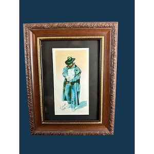 May include: A framed print featuring a drawing of a figure wearing a cowboy hat, a long coat, and jeans. The artwork is signed by the artist and set within a dark brown wooden frame with ornate carved details and a gold-toned inner border.