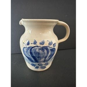 May include: Cream-colored ceramic pitcher with a blue apple design. The pitcher has a curved handle and a spout for pouring. The apple design is hand-painted with leaves and stems. A decorative piece for the kitchen or dining room.