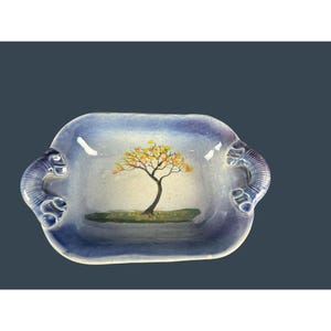 Puede incluir: Una fuente rectangular de cerámica con un degradado azul y blanco. La fuente presenta un árbol pintado con hojas amarillas y naranjas. Las asas son de un azul más oscuro con un diseño de remolino decorativo.