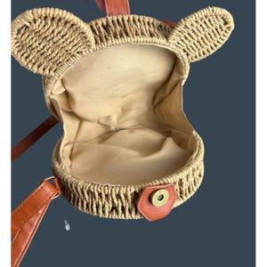 May include: A round, woven straw handbag with bear ears. The interior is lined with beige fabric. It has a brown leather strap and a snap closure. The bag is designed to be carried over the shoulder.