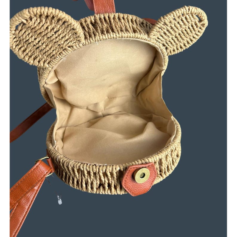 May include: A round, woven straw handbag with bear ears. The interior is lined with beige fabric. It has a brown leather strap and a snap closure. The bag is designed to be carried over the shoulder.