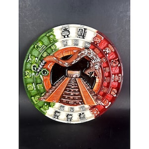 Könnte beinhalten: Ein runder Wandbehang aus Keramik mit einem farbenfrohen Design, das den aztekischen Kalender darstellt. Der Kalender ist in den Farben Grün, Weiß und Rot gehalten, mit einer Schlange, die sich um die Mitte windet. Die Mitte des Kalenders zeigt eine Pyramide mit Stufen.