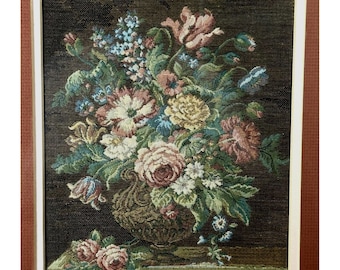May include: A framed floral tapestry featuring a detailed arrangement of colorful flowers in a decorative vase. The flowers include pink, yellow, blue, and white blooms with green foliage. The tapestry is set against a dark background and framed in white.