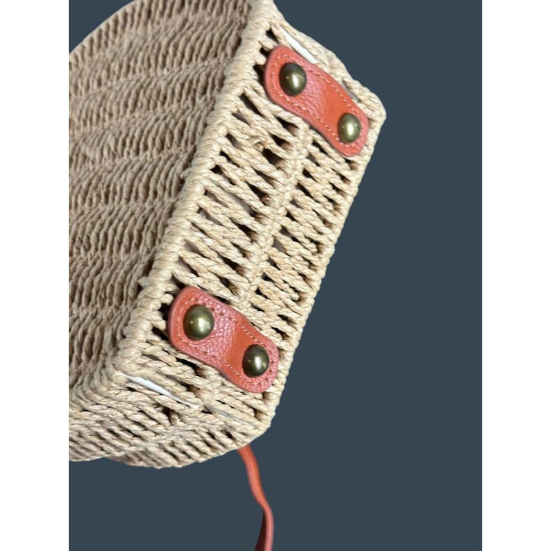 May include: A woven straw handbag with a rectangular shape. The bag features tan leather accents and brass-colored rivets. The bag has a woven texture and a handle made of the same material. The bag is designed for carrying items.