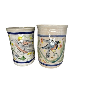 May include: Two ceramic mugs with hand-painted floral designs and birds. One mug features a blue bird with a yellow beak and the other mug features a pink bird with a yellow beak.