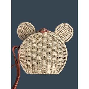 May include: A woven straw crossbody bag in the shape of a mouse, with two circular ears and a round body. The bag has a brown leather strap and a small leather accent on top. The bag is tan and brown.